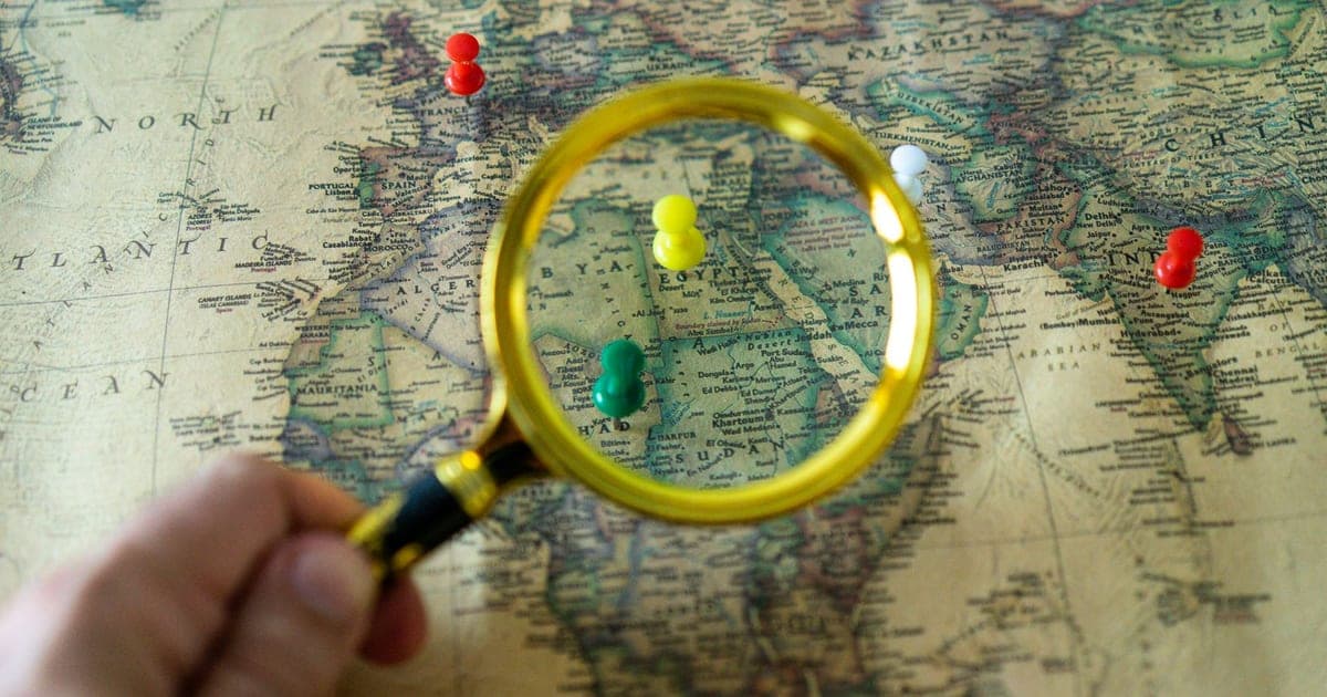 Una mano tiene una lente d'ingrandimento sopra una mappa del mondo d'epoca, con puntine colorate in Nord Africa, in Europa e in India