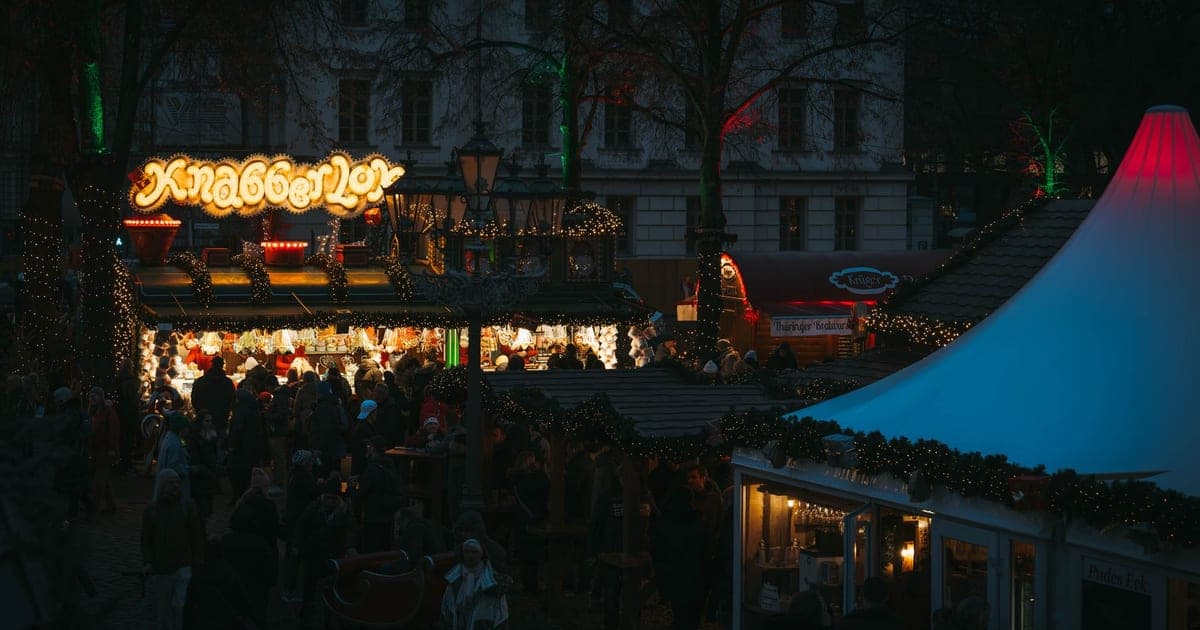 Wooden market stalls lit with warm lights in front of a Gothic cathedral at dusk, steam rising from mulled wine cups