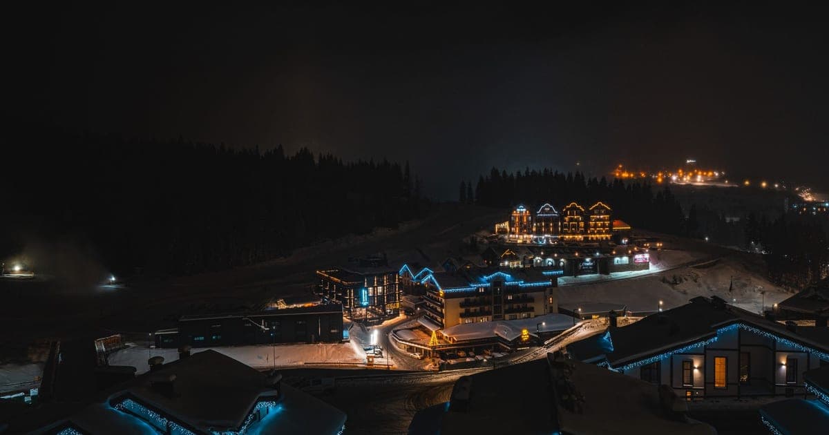 A snow-covered ski village at night, with lit chalets and hotels below a dark forest